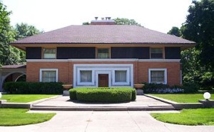 Winslow House: A house with a rectangular base and triangular roof with two distinct floors, a white front doorway with a dark door, and a two-toned brick facade. 