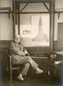 August Perret (1874,1954), foto d'epoca: Una foto in bianco e nero di Perret con le gambe incrociate e un poster di un grande edificio. 