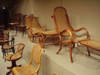 A photo of Thonet’s bentwood chairs in the setting of a warehouse. The chairs are made of a light-colored wood and a weaved seat and backing.