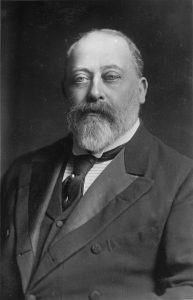 Photo of Edward VII: A large white man with a full beard, yet thinning hair upon his hair. He is dressed in a dark suit. 
