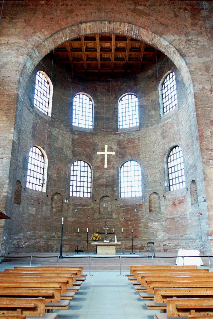 Photo of the inside of the Basilica of Constantine. A circular dome-like structure extends onto a rectangular room. There are church pews on the bottom right and left of the photo, with an isle in the center. Moreover, there is an altar and the end of the isle and a white cross sitting above the altar. 