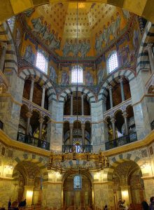 Cappella Palatina, Aquisgrana. Una foto del soffitto della cupola nella cappella. Ci sono due file di archi con mini finestre ad arco in cima. Inoltre, la parte superiore del soffitto è in oro con varie immagini religiose.