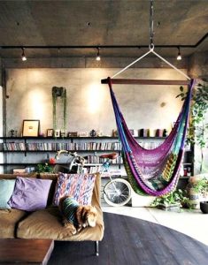Example of a Boho interior, with a hammock and a bike leaned on a bookcase. There is also a brown couch with bright colored pillows on it. 