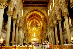 Vista interna della Cappella Palatina situata al piano terra al centro del Palazzo Reale di Palermo, Italia meridionale. Le colonne si ergono su entrambi i lati della navata e una luce luminosa e dorata occupa lo sfondo, al centro del Palazzo. 
