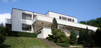 Villa Tugendhat pictured trom the exterior. It is a large white building that sits elevated. 