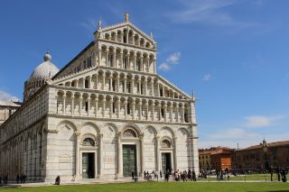 Catedral de Pisa em estilo românico. Um grande relvado estende-se em frente à catedral, com dezenas de turistas à espera na fila para entrar. 