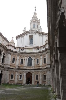 Santo Ivo alla Sapienza em Roma, estilo barroco romano. O pátio de uma estrutura branca em forma de castelo. Tem dois níveis com janelas em arco colocadas simetricamente em toda a sua extensão. Além disso, uma torre pontiaguda ergue-se no centro da imagem. 