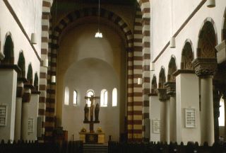 Igreja de São Miguel, Hildesheim, Alemanha. Uma igreja simples com arcos estriados, paredes brancas e castanhas e um crucifixo no fim da nave. Pequenas janelas em arco estão situadas no meio da cúpula, no fundo da fotografia.