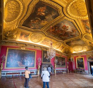 Salão de Marte no Palácio de Versalhes ao estilo Luís XIV. Uma grande sala com paredes vermelhas, grandes pinturas intrincadas e um tecto dourado com três grandes pinturas.