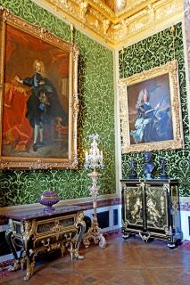 Mobiliário de estilo Luís XIV. Uma sala com paredes verdes, duas molduras douradas com grandes retratos e mobiliário escuro com apontamentos dourados. Além disso, o tecto parece ser banhado a ouro.