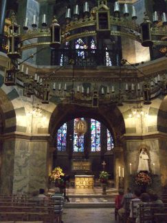 Interior da Capela Palatina na Catedral de Aachen, na Alemanha. Uma grande sala com um lustre suspenso, uma janela com vitrinas e vários elementos religiosos. Além disso, há convidados nos bancos em primeiro plano da fotografia. 