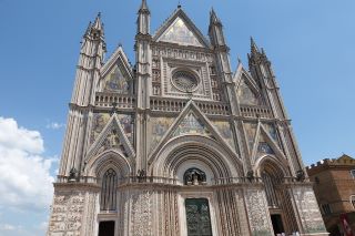 Fachada da Catedral de Orvieto - estilo gótico do século XIV. Uma grande estrutura de pedra cinzenta decorada com vários arcos e pontos triangulares. 