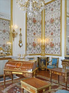 Mobiliário de estilo Luís XIV. Uma grande sala com paredes brancas e móveis de madeira com detalhes dourados ocupam o quarto.