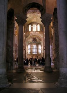 San Vitale a Ravenna. Ci sono due colonne in primo piano, con la zona della cupola occupata da turisti e grandi finestre ad arco. Inoltre, la cupola è coperta di intricati dipinti religiosi. 