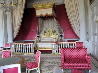 Quarto Luís XIV com mobiliário de bolinhas cor-de-rosa e uma grande cama branca com detalhes dourados.  