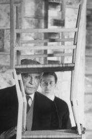 Gio Ponti, ritratto in bianco e nero, mentre guarda attraverso lo spazio fra due sedie.