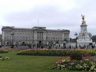 O Palácio de Buckingham e a estátua em memória da Rainha Vitória. O Palácio de Buckingham está ao fundo, com um grande jardim com flores vermelhas e amarelas em primeiro plano. 