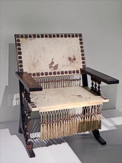 Fauteuil di Carlo Bugatti. Una sedia in legno scuro con schienale e seduta leggeri. Inoltre, le nappe pendono dalla parte inferiore dello schienale e dalla parte anteriore del sedile. 
