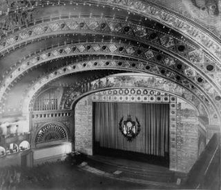 Fotografia do Auditorium Theatre visto do interior a partir do balcão, Chicago. Pode ver-se na fotografia um grande tecto que se encontra por cima do palco e várias pessoas na plateia por baixo. 
