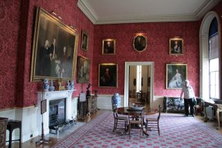 Um exemplo do estilo Regency numa sala de paredes vermelhas com vários retratos emoldurados a ouro e uma pequena mesa de madeira no centro. 