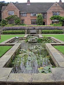 Goddards House and Garden, York, Inghilterra. Un grande stagno con vegetazione occupa il primo piano della foto, mentre una grande casa con finestre rettangolari occupa lo sfondo. 