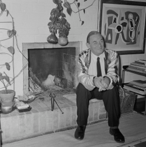 Alvar Aalto photo from 1956 in black and white. He sits on the bottom of a fireplace. 