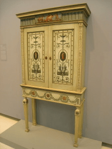 A yellow-toned bookcase with two doors featured with painted, floral designs and four skinny legs along the bottom. 
