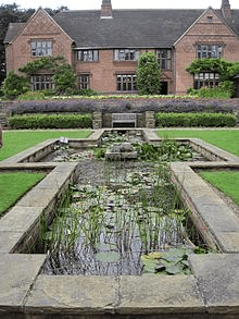 Goddards House and Garden, York, England. A large pond with plants is in the foreground of the photo, while the background is covered with a big house with rectangular windows displaced on the front of the house. 