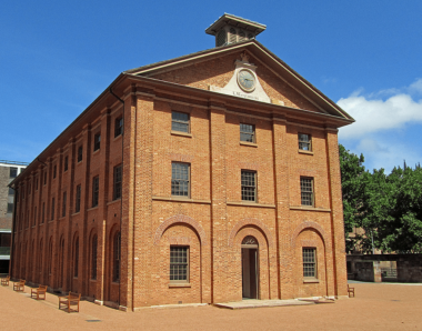 Hyde Park Barracks, which is a brown-colored building; the facade shows three floors, each with three small rectangular windows. 