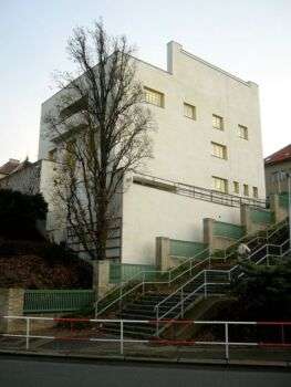 Adolf Loos- Muller House, 1929-30: A photo of the side of the house, where there is a green-fence along the stairs. Moreover, the building is a big white structure with yellow windows. 