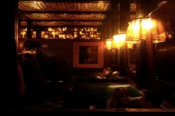 American Bar, Vienne, Autriche, 1907, Adolf Loos : Un bar sombre éclairé avec des lampes aux tons chauds le long du mur droit et des éléments simples et romantiques dans tout l'espace.