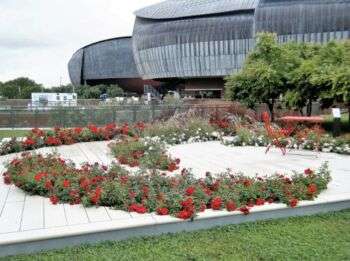 Photo of the Auditorium Parco della Musica, Roma, from the outside. On the foreground, some bushes arranged in a circle, with blossomed red flowers in it. 
