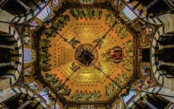 Capela do Palatino em Aachen, pormenor do mosaico da cúpula bizantina. Uma cúpula octogonal dourada exposta a partir de baixo. Há várias pessoas pintadas ao longo dos bordos. Além disso, os vértices dos arcos que suportam a cúpula alinham-se com a moldura do quadro.
