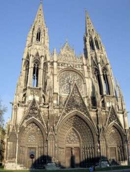 The Church of St. Ouen, Rouen, Normandy- France. It is a large tan-colored cathedral with sharp points and decorations pointing towards the sky. 