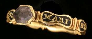 Exemplo de um anel modernista com uma pedra hexagonal escura e o nome "Mary" gravado na parte circular.