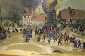 Franco-Prussian war painting. There are two houses in the background and blue soldiers in the front. 