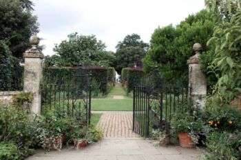 Hidcote Manor Garden: A photo of the entrance with a black metal gate with two stone posts on either side. Moreover, the garden ahead has tall bushes and a stone path. 
