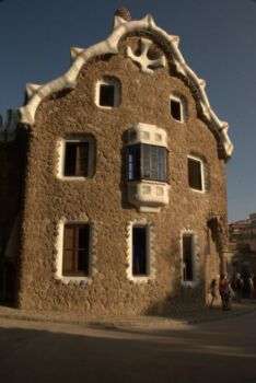 A photo of a house in Park Guell,  a large medium-brown structure with a while roof and carved out rectangular windows. 
