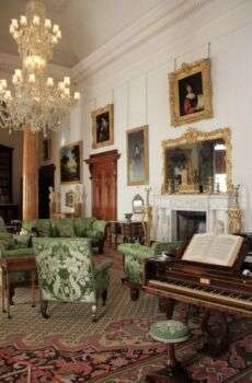 A room decorated in the Regency style. There is an ornate chandelier in the center, various gold-framed portraits, green floral furniture, a white fireplace and a medium-wood piano. 