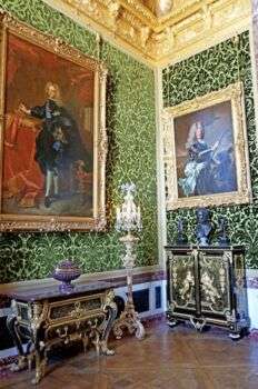 A room with green-colored walls, two gold picture frames with grand portraits, and dark furniture with gold accents. Moreover, the ceiling appear to be gold accented as well. 