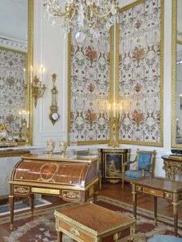 A large room with white walls, and wooden furniture with gold accents that occupy the room. 