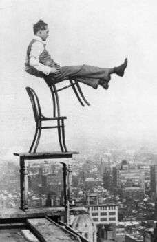 Michael Thonet chair balance, photo of a man on a chair balancing on another chair, which is on a table on the edge of a skyscraper. 