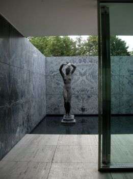 Mies van der Rohe Pavilion. Detail of a statue of a naked woman, realized in a dark opaque material. The woman has both of her hands above her head.  