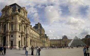 A photo of the Louvre, on the right there is the glass pyramid, and there are tourists all around in the photo. 