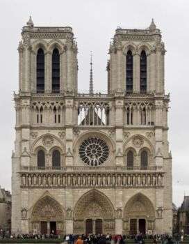 Photo of Notre Dame from the outside. Two towers sit side by side with various ornate architectural details. There are three arched doorways along the ground, the second row has a circle center, with two side by side arches on either side, as well as a pointed tower in the center. 