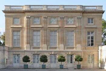 Le Petit Trianon: A light tan-stoned building with large rectangular windows, four trees along the front a 5 sets of double doors along the ground. 