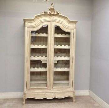 Armario del estilo Rococó transformado en una pequeña bodega de 44 botellas. Un armario de madera clara con dos puertas de vidrio. Los bordes tienen florituras sinuosas y hay estantes de vino al centro de la pieza. 
