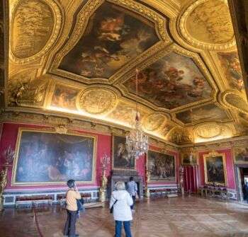A grand room with red walls, large intricate paintings and a gold ceiling with three grand paintings. 