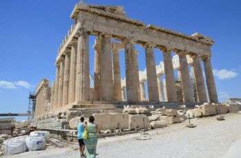 Photo of the Parthenon with blue, clear skies. Two people stand in the bottom left hand corner. 