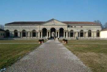 Photo of the palazzo Te Mantova, which is a long building with many arches. Three arches occupy the center of the photo with a gravel path surrounded by grass leading up to it. Moreover, a blue sky occupies the background. 